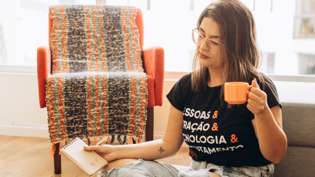 Mulher sentada no chão segurando um caderno da Singularis RH em uma mão, uma xícara de café em outra e um notebook em sua frente