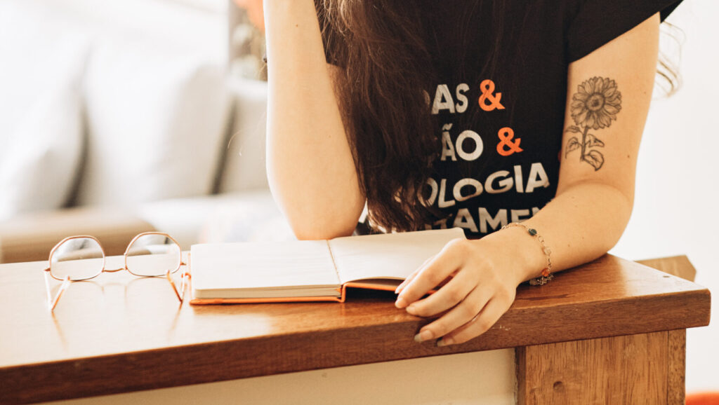 Uma mulher apoiada sobre uma bancada com um caderno da Singularis RH em sua frente e um óculos ao lado do caderno pensando em uma descrição de vaga