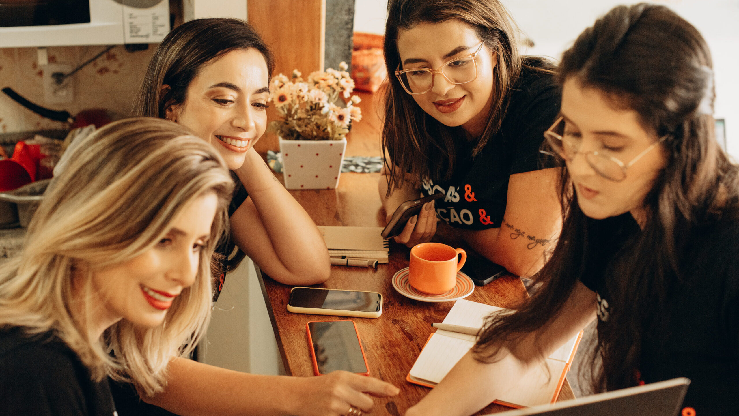 Grupo de quatro mulheres sorridentes reunidas em volta de uma mesa, trabalhando em um laptop e discutindo ideias, com cadernos e celulares à vista, em um ambiente descontraído e acolhedor.