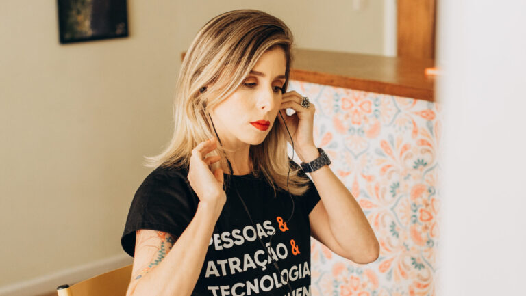 Mulher usando uma camiseta preta com as palavras 'Pessoas, Atração, Tecnologia, Recrutamento' e o logo da Singularis, colocando fones de ouvido enquanto se prepara para uma videoconferência em um ambiente acolhedor com uma xícara de café e um laptop sobre a mesa.