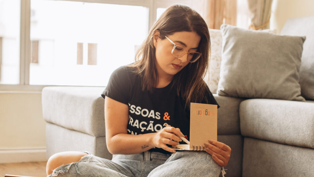 Mulher sentada no chão de uma sala, com um notebook no colo e uma xícara de café ao lado, escrevendo em um caderno com concentração, em um ambiente confortável e iluminado.