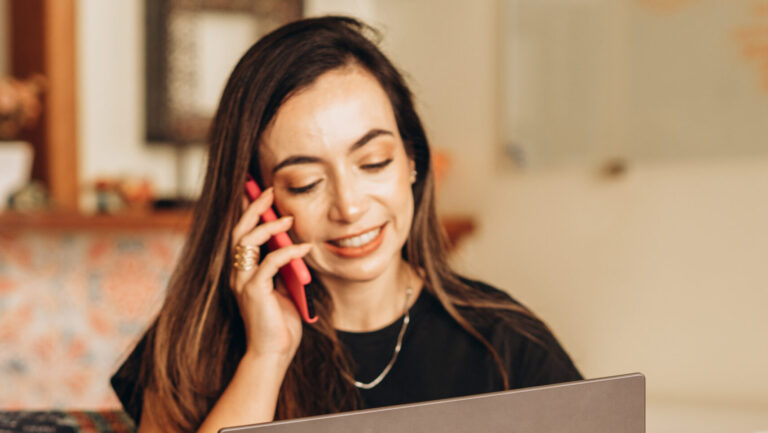 Mulher sorridente sentada em uma poltrona vermelha, falando ao telefone enquanto utiliza um laptop Asus em seu colo, em um ambiente acolhedor com decoração colorida ao fundo.