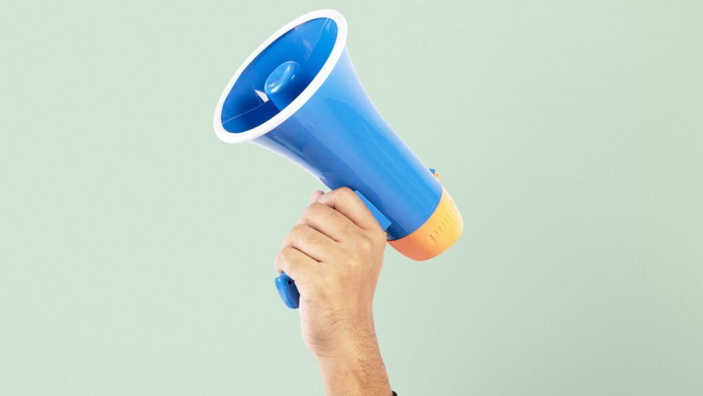 Mão segurando um megafone azul com detalhes em laranja, levantado contra um fundo verde claro, simbolizando uma etapa do processo de recrutamento e seleção.