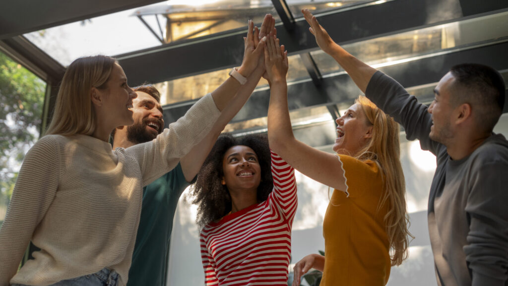 Grupo de cinco colegas de trabalho celebrando com um high-five coletivo, sorrindo e mostrando entusiasmo em um ambiente iluminado com janelas amplas.