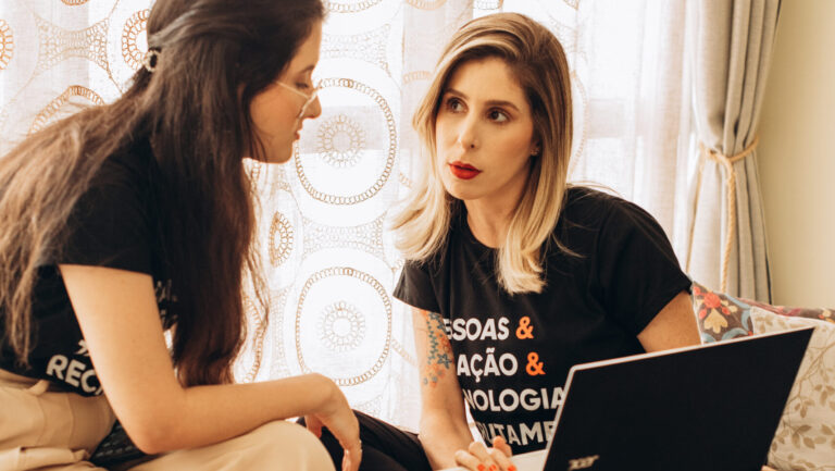 Duas mulheres conversando, uma delas com um laptop no colo, em um ambiente acolhedor e iluminado. Ambas vestem camisetas com palavras relacionadas a pessoas, tecnologia e recrutamento.