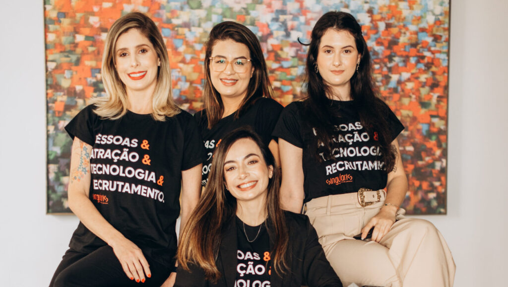 Equipe de quatro mulheres posando em ambiente interno, usando camisetas pretas com dizeres relacionados a recrutamento e tecnologia, com um quadro colorido ao fundo.