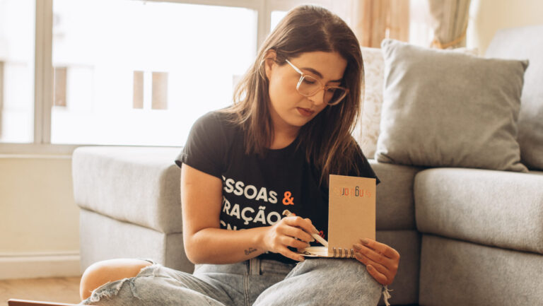 Mulher sentada no chão de uma sala iluminada, vestindo óculos e uma camiseta preta, escrevendo em um caderno enquanto olha para ele.