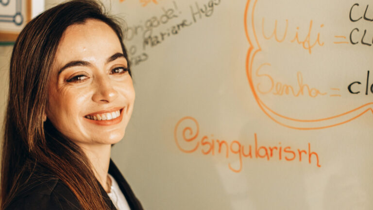 Mulher sorridente em frente a um quadro branco, segurando um marcador vermelho, com textos e desenhos escritos ao fundo, incluindo o handle '@singularisrh'.