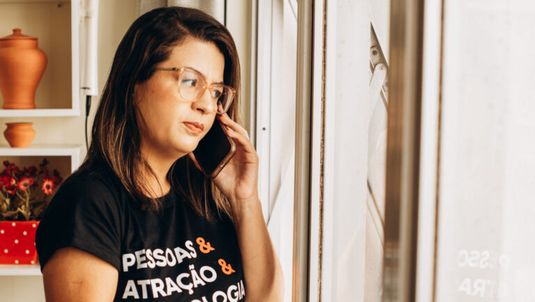 Mulher com óculos e cabelos longos usando uma camiseta preta com os dizeres "PESSOAS & ATRAÇÃO & TECNOLOGIA & RECRUTAMENTO" enquanto fala ao telefone, olhando pensativa pela janela. Ela está próxima a uma estante branca com vasos decorativos e flores.