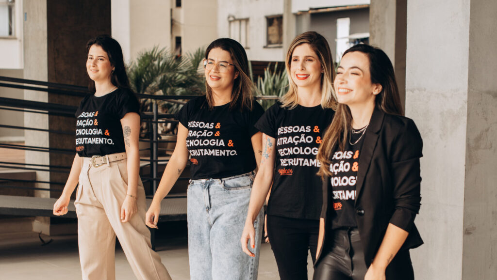Um grupo de quatro mulheres caminha lado a lado em um ambiente moderno. Elas vestem camisetas pretas com dizeres sobre "PESSOAS & ATRAÇÃO & TECNOLOGIA & RECRUTAMENTO", combinadas com diferentes estilos de calças e calçados. Elas sorriem e aparentam estar em um momento descontraído, com uma vibe profissional e confiante.