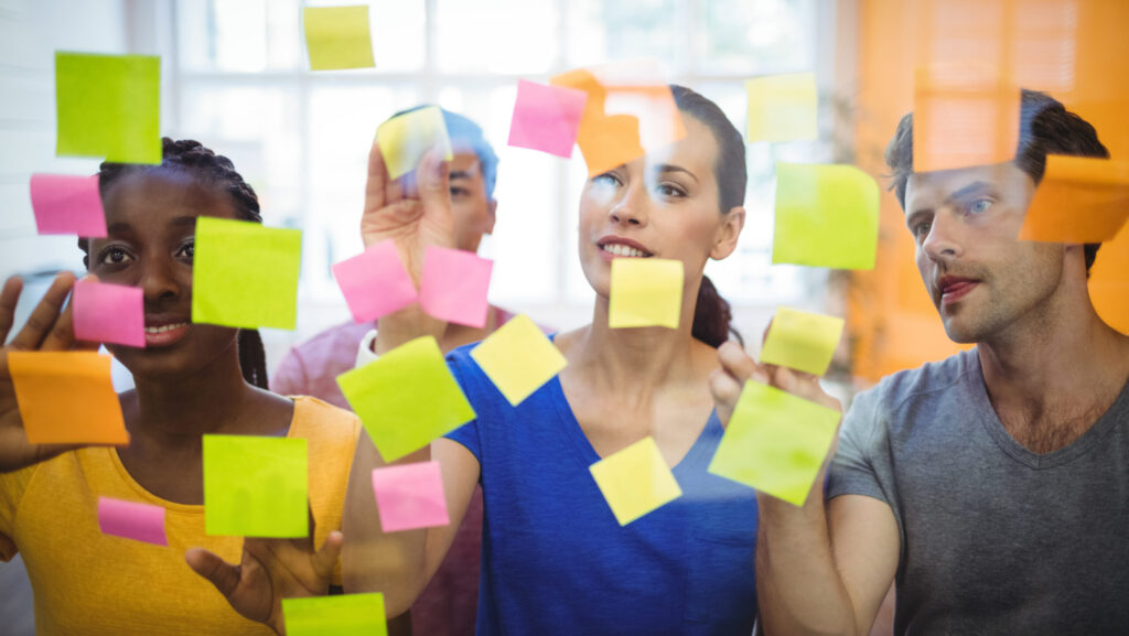 Grupo de profissionais colando e organizando notas adesivas coloridas em um painel de vidro, colaborando em uma atividade de brainstorming, com um ambiente iluminado ao fundo.