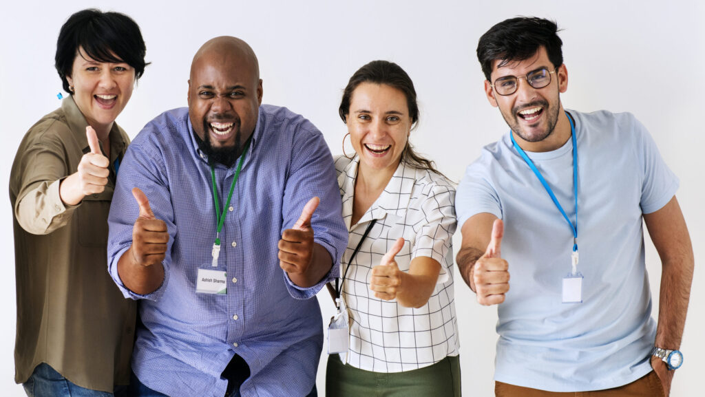 Grupo diverso de colegas de trabalho sorrindo e mostrando aprovação com polegares para cima, representando engajamento e motivação no ambiente corporativo.