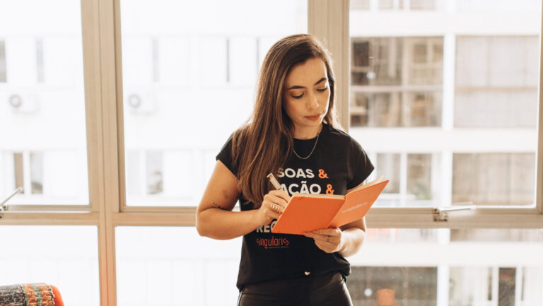 Profissional da Singularis RH segurando caderno laranja enquanto faz anotações em um ambiente iluminado, com cadeira vermelha e xícara laranja sobre um apoio ao fundo.