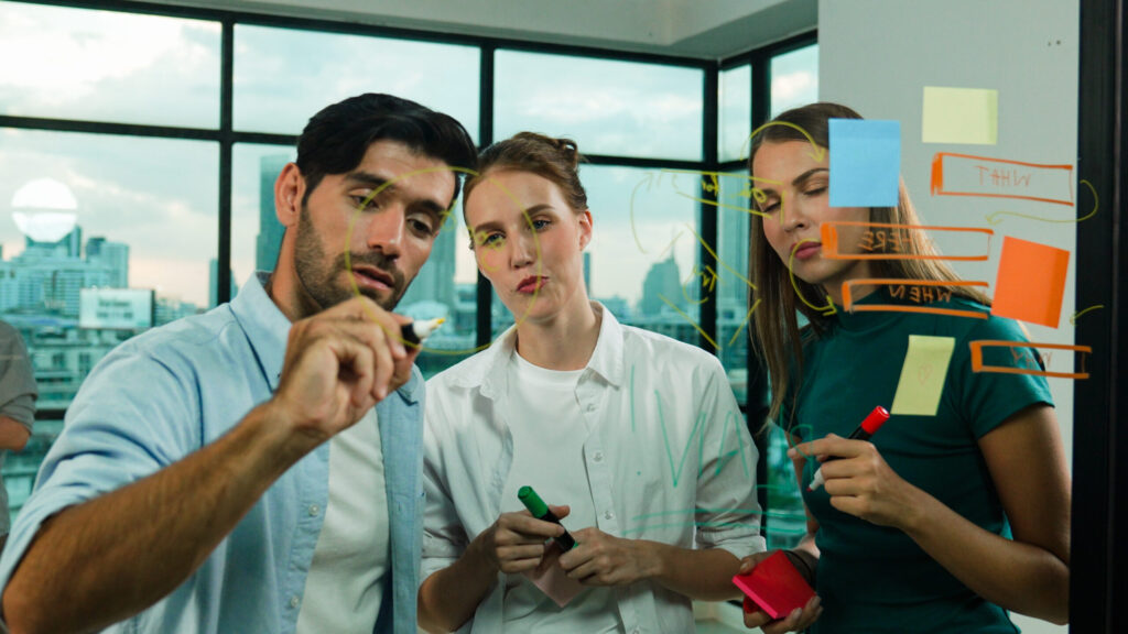 Equipe de trabalho colaborando em frente a um quadro de vidro com anotações e post-its, enquanto discutem estratégias em um escritório moderno com vista para a cidade.