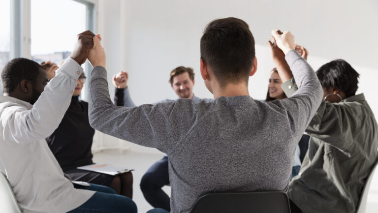 Grupo de pessoas sentadas em círculo, segurando as mãos para cima, demonstrando união, trabalho em equipe e retenção de talentos, em uma sala bem iluminada.