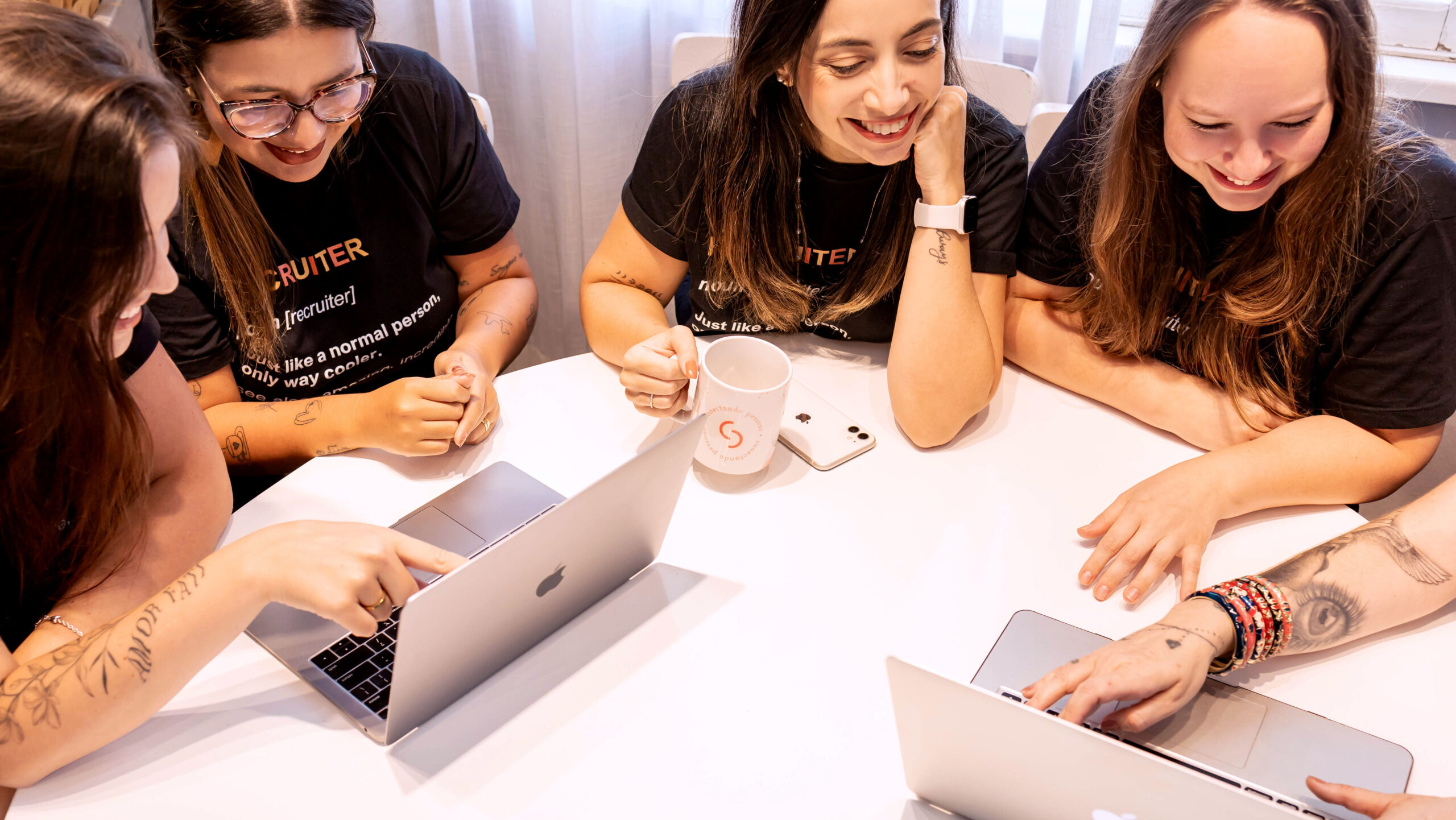 Grupo de mulheres sorrindo e colaborando em torno de uma mesa com laptops, enquanto compartilham ideias e interagem em um ambiente descontraído.