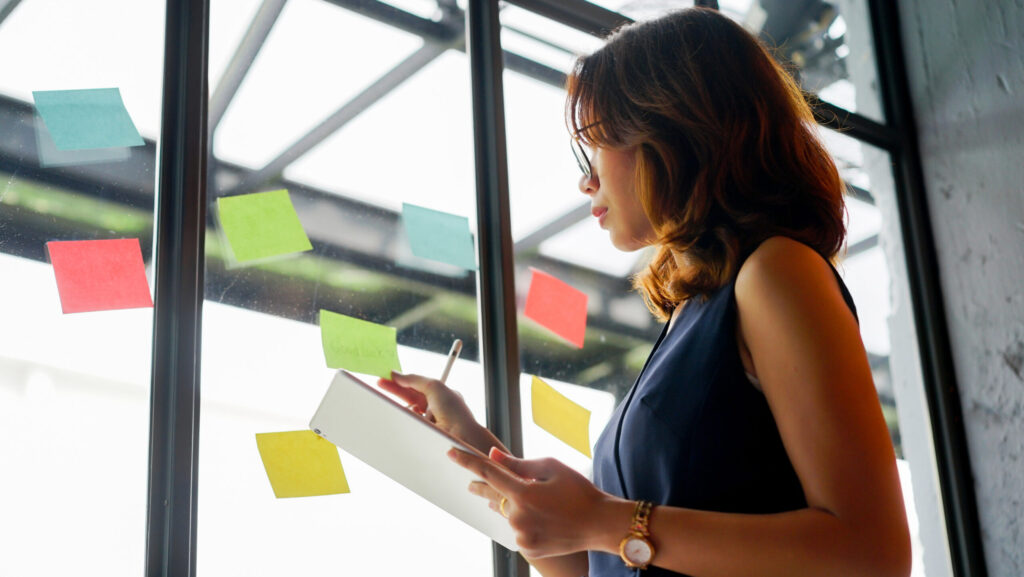 Mulher realizando onboarding de colaboradores, segurando um bloco de notas em frente a uma janela com post-its coloridos, representando organização e integração de novos colaboradores em empresas de tecnologia.